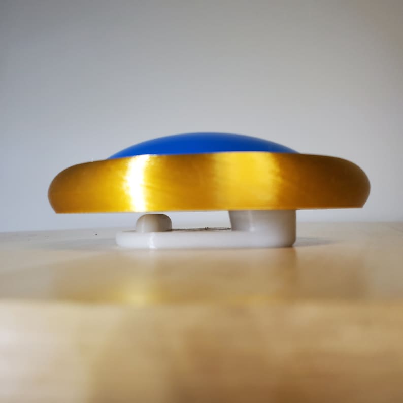May include: A blue and gold button with a white base. The button is round and has a shiny gold rim.