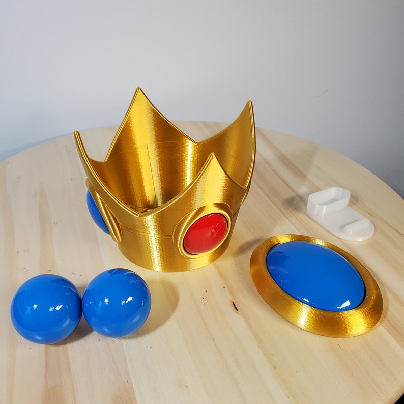 May include: A gold crown with red and blue gems, two blue spheres, and a gold disc with a blue center. The crown is a 3D printed object.