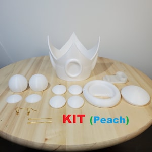 May include: A white 3D printed Princess Peach cosplay kit. The kit includes a crown, a headband, two earrings, two hair clips, and eight small round pieces. The text "KIT (Peach)" is written on a wooden surface.