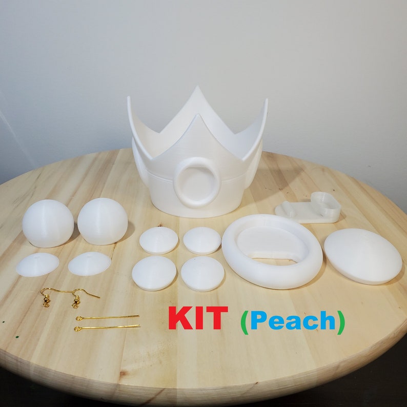 May include: A white 3D printed Princess Peach cosplay kit. The kit includes a crown, a headband, two earrings, two hair clips, and eight small round pieces. The text "KIT (Peach)" is written on a wooden surface.