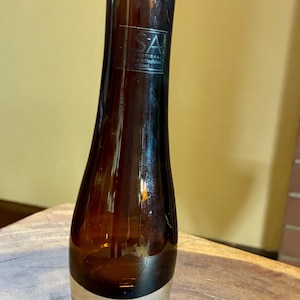 May include: A brown glass vase with a unique, curved shape. The vase has a clear glass base and a dark brown body. The top of the vase is slightly wider than the base. The vase has a label that reads "SA International".
