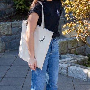 K&ouml;nnte beinhalten: Eine Person tr&auml;gt ein schwarzes T-Shirt mit einer wei&szlig;en Tragetasche, die ein kleines Blumenmuster aufweist. Die Person tr&auml;gt auch blaue Jeans und wei&szlig;e Turnschuhe.