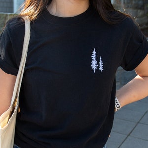 K&ouml;nnte beinhalten: Ein schwarzes T-Shirt mit einem wei&szlig;en gestickten Design aus zwei B&auml;umen auf der linken Brust.