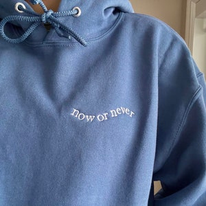 May include: A dark blue hooded sweatshirt with the phrase "now or never" embroidered in white on the front.