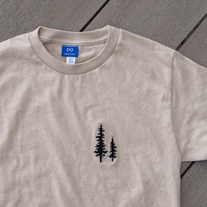 K&ouml;nnte beinhalten: Ein beiges T-Shirt mit einem kleinen gestickten Design aus zwei Kiefern auf der linken Brust.