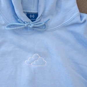 May include: A light blue hoodie with a white embroidered cloud design. The hoodie has a drawstring hood and a small tag on the front that says "(c) DREAM".