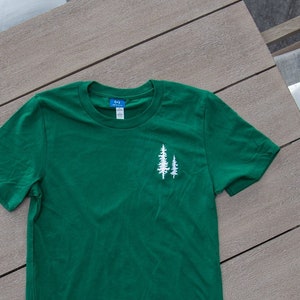 K&ouml;nnte beinhalten: Ein gr&uuml;nes T-Shirt mit einer wei&szlig;en Grafik von zwei B&auml;umen auf der linken Brust.