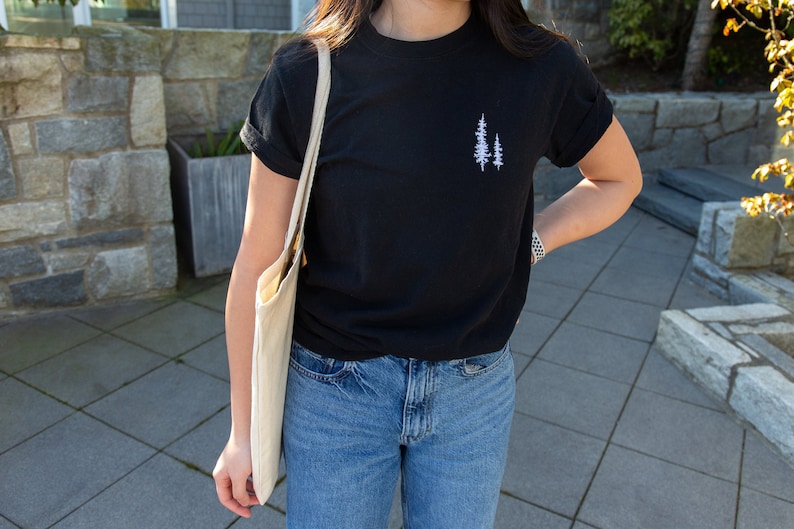 K&ouml;nnte beinhalten: Ein schwarzes T-Shirt mit einem wei&szlig;en gestickten Design aus zwei B&auml;umen auf der linken Brust.