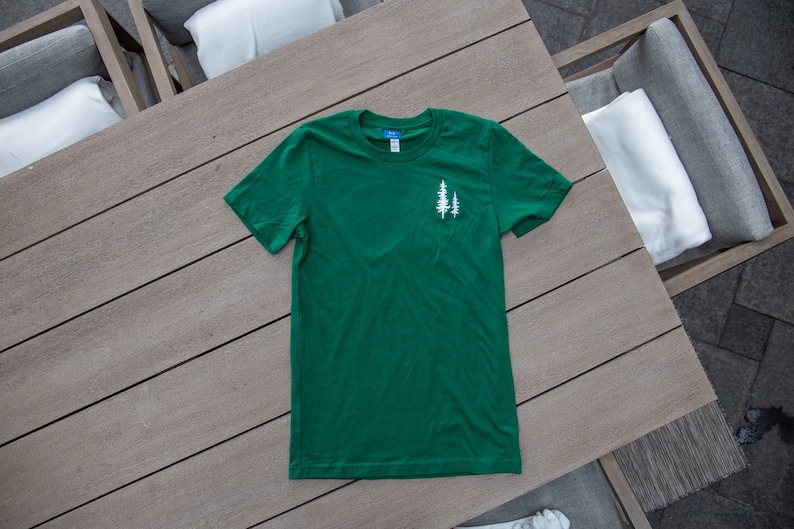 K&ouml;nnte beinhalten: Ein gr&uuml;nes T-Shirt mit einer wei&szlig;en Grafik von zwei B&auml;umen auf der linken Brust.