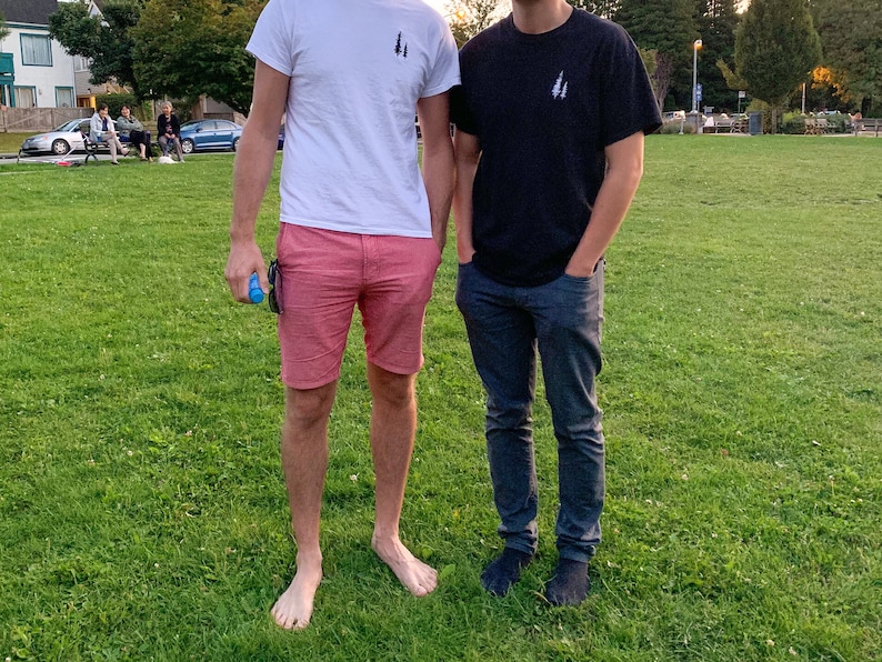 K&ouml;nnte beinhalten: Zwei Personen stehen auf einem grasbewachsenen Feld und tragen T-Shirts mit einem kleinen Baummotiv auf der Vorderseite. Die Person links tr&auml;gt eine rosa Shorts und die Person rechts tr&auml;gt eine blaue Jeans.