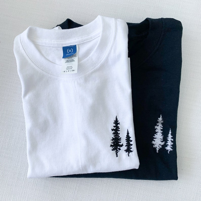 K&ouml;nnte beinhalten: Zwei T-Shirts, eines wei&szlig; und eines schwarz, mit einem gestickten Design aus drei Kiefern auf der linken Seite jedes Shirts.