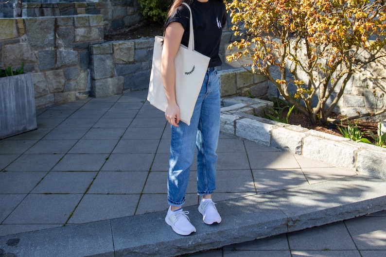 K&ouml;nnte beinhalten: Eine Person tr&auml;gt ein schwarzes T-Shirt mit einer wei&szlig;en Tragetasche, die ein kleines Blumenmuster aufweist. Die Person tr&auml;gt auch blaue Jeans und wei&szlig;e Turnschuhe.