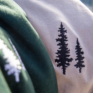 K&ouml;nnte beinhalten: Zwei T-Shirts in verschiedenen Farben mit gestickten Kiefernbaum-Designs. Ein Shirt ist gr&uuml;n und das andere beige.