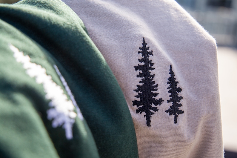K&ouml;nnte beinhalten: Zwei T-Shirts in verschiedenen Farben mit gestickten Kiefernbaum-Designs. Ein Shirt ist gr&uuml;n und das andere beige.