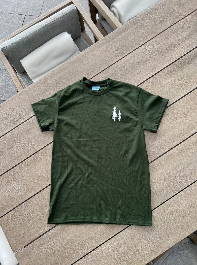 K&ouml;nnte beinhalten: Ein dunkelgr&uuml;nes T-Shirt mit einem wei&szlig;en Umriss von zwei Kiefern auf der Vorderseite.