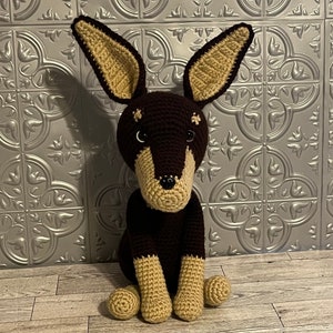 May include: A crocheted brown and tan dog toy with black eyes and a black nose. The toy is sitting on a wooden floor.