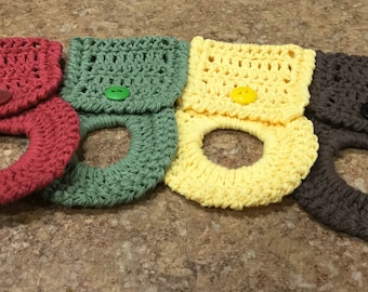 May include: Four handmade crochet towel holders in shades of red, green, yellow, and brown. Each holder features a button closure and a circular opening for a towel. The crochet work is visible, showcasing the texture and craftsmanship.