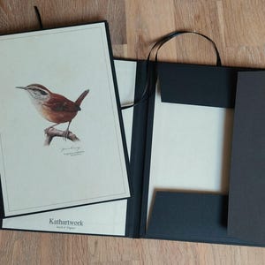 Könnte beinhalten: Ein schwarzer Ordner mit einem braunen Papiereinsatz, der eine Aquarell-Illustration eines braunen Zaunkönigs zeigt, der auf einem Ast sitzt. Der Ordner hat eine Klappe mit einem schwarzen Papiereinsatz und einem schwarzen Bandverschluss. Der Text "Kathartwork" ist auf der Unterseite des Ordners gedruckt.