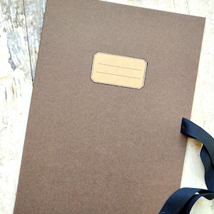 Könnte beinhalten: Ein brauner Papierordner mit einem leeren Etikett und einer schwarzen Schleife. Der Ordner liegt auf einem hellen Holzuntergrund.