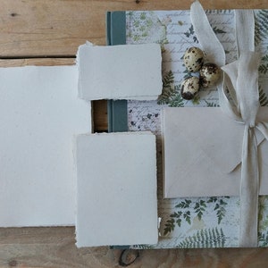 Puede incluir: Un juego de papeler&iacute;a con papel hecho a mano, un sobre y un diario con un dise&ntilde;o de helechos y escritura. El conjunto est&aacute; atado con una cinta de color claro e incluye tres huevos de codorniz moteados. El papel tiene un borde irregular.