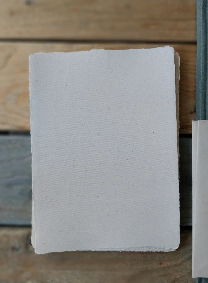 Puede incluir: Una pila de papel hecho a mano de color blanquecino con bordes irregulares. El papel tiene una superficie ligeramente texturizada y es de forma rectangular. El papel es probablemente para arte o escritura. Un lomo de libro verde azulado es visible a la derecha.