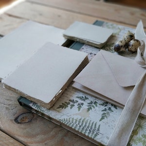 Puede incluir: Una colecci&oacute;n de art&iacute;culos de papeler&iacute;a, que incluye pilas de papel blanquecino, sobres y un cuaderno con una cubierta estampada con helechos. Una cinta beige est&aacute; atada en un lazo. Los art&iacute;culos est&aacute;n dispuestos sobre una superficie de madera r&uacute;stica.
