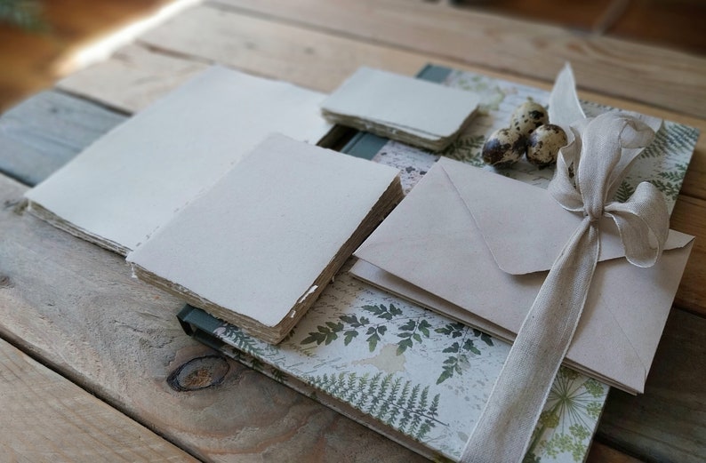 Puede incluir: Una colecci&oacute;n de art&iacute;culos de papeler&iacute;a, que incluye pilas de papel blanquecino, sobres y un cuaderno con una cubierta estampada con helechos. Una cinta beige est&aacute; atada en un lazo. Los art&iacute;culos est&aacute;n dispuestos sobre una superficie de madera r&uacute;stica.