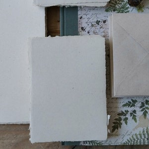 Puede incluir: Colecci&oacute;n de productos de papel hechos a mano, incluyendo hojas rectangulares y cuadradas con bordes irregulares, un sobre y un peque&ntilde;o libro. El papel es blanquecino, con un fondo de papel estampado con elementos bot&aacute;nicos y de escritura.