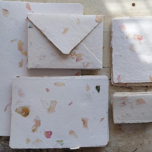 Könnte beinhalten: Ein Set aus handgeschöpftem, cremeweißem Briefpapier mit eingearbeiteten getrockneten Blütenblättern in Rosa, Gelb und Grün. Das Set enthält verschiedene rechteckige Bögen, einen gefalteten Umschlag und kleine Notizkarten mit Büttenrand.