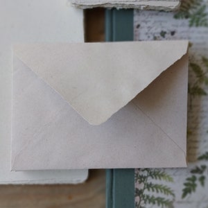 Puede incluir: Un peque&ntilde;o sobre blanquecino con una superficie texturizada. El sobre es rectangular con una solapa triangular. El papel tiene un aspecto ligeramente &aacute;spero, hecho a mano. El fondo incluye otros elementos de papel y bot&aacute;nicos.