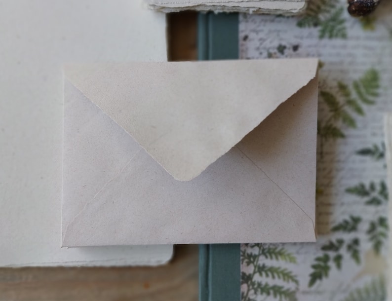 Puede incluir: Un peque&ntilde;o sobre blanquecino con una superficie texturizada. El sobre es rectangular con una solapa triangular. El papel tiene un aspecto ligeramente &aacute;spero, hecho a mano. El fondo incluye otros elementos de papel y bot&aacute;nicos.