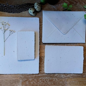Könnte beinhalten: Ein Set aus hellgrauem, handgeschöpftem Briefpapier, bestehend aus einem rechteckigen Bogen, einer kleineren rechteckigen Karte und einem passenden Umschlag. Das Papier hat eine texturierte, gesprenkelte Oberfläche mit unebenen Büttenrändern.