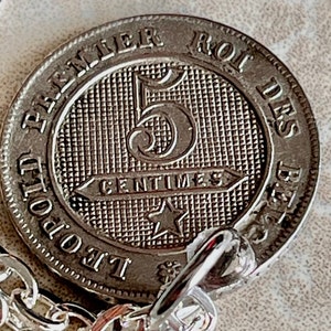 May include: A silver coin with the inscription "PREMIER ROI DES BELGES" and "5 CENTIMES" around a large number "5" with a star below. The coin is attached to a silver chain.