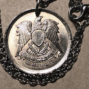 Puede incluir: Collar de cadena de plata con un colgante de moneda de plata que presenta el escudo de armas de los Emiratos Árabes Unidos, que incluye un águila con un escudo y una pancarta con texto árabe.