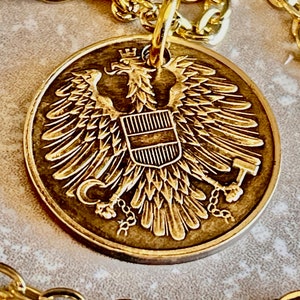 May include: A gold-colored pendant featuring a detailed image of a heraldic eagle with a shield on its chest. The eagle is surrounded by a chain.