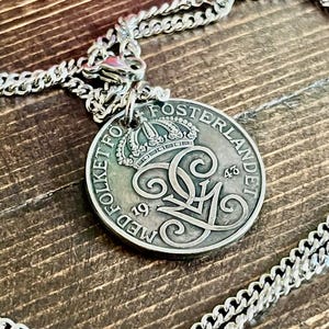 Puede incluir: Colgante de moneda plateada con un diseño detallado, que incluye una corona y letras ornamentadas. La moneda está unida a una cadena plateada. La moneda dice "MED FOLKET FOR FOSTERLANDET" y tiene los números "19" y "43".
