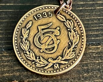 Könnte beinhalten: Ein kreisförmiger Anhänger in Bronzetönen mit einem Vintage-Münzdesign aus dem Jahr 1935. In der Mitte befindet sich eine stilisierte 5 mit einem Schrägstrich, umgeben von einem geprägten Lorbeerkranz, der unten mit einer Schleife gebunden ist.