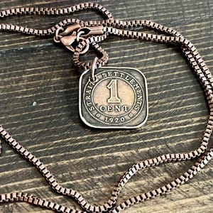 May include: A copper-toned necklace with a square pendant. The pendant displays the text "STRAIT SETTLEMENTS 1 CENT 1920". The chain and pendant are copper-colored. The necklace is on a wooden surface.