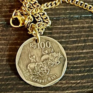 May include: Gold-colored chain necklace with a round pendant. The pendant features an image of people riding animals and the text "Rp 100" and "Karapan Sapi". The necklace is displayed on a dark wooden surface.