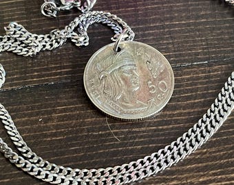Könnte beinhalten: Ein silberfarbener Münzanhänger an einer passenden Panzerkette. Die Münze zeigt ein Profilporträt einer Person mit Stirnband, die Zahl 50 und das eingeprägte Jahr 1950. Ein kleiner Metallring verbindet die Münze mit der Kette.