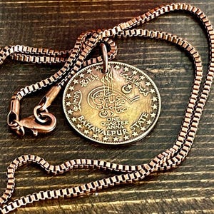 May include: Copper-toned necklace featuring a round pendant with ornate text and star accents. The pendant reads "ONE QUARTER ANNA BAHAWALPUR STATE". The chain is a box link style with a lobster clasp.