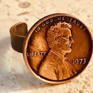 May include: A copper-colored ring made from a 1973 penny. The penny features the profile of a man and the words "IN GOD WE TRUST" and "LIBERTY". The ring band is a simple, rounded design.