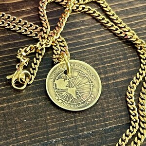 May include: A gold-coloured necklace with a round pendant featuring a map and the text "REPUBLICA DEL ECUADOR LUZ DE AMERICA". The chain is a thick curb link style, and the clasp is visible. The necklace is displayed on a dark wooden surface.