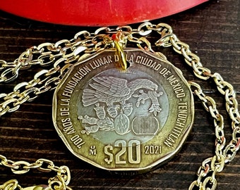 Könnte beinhalten: Eine goldfarbene Metallkettenhalskette mit einem runden 20-Peso-Münzanhänger. Die Münze zeigt einen Adler auf einem Kaktus sowie den Text 700 AÑOS DE LA FUNDACIÓN LUNAR DE LA CIUDAD DE MÉXICO - TENOCHTITLAN, $20 und 2021.