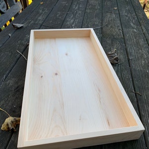 May include: A rectangular wooden tray with a natural wood finish. The tray is empty and has a raised edge.
