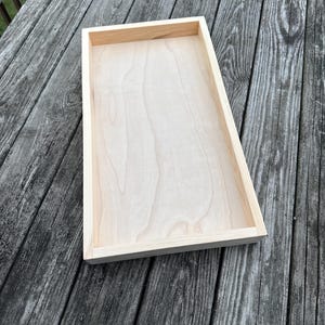 May include: A rectangular wooden tray with a light-colored, unfinished wood surface and raised edges. The tray is empty and sits on a weathered wooden deck. The tray's dimensions are not visible in the image.
