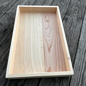 May include: A rectangular wooden tray with a natural wood finish. The tray has a raised edge and is made of two pieces of wood.