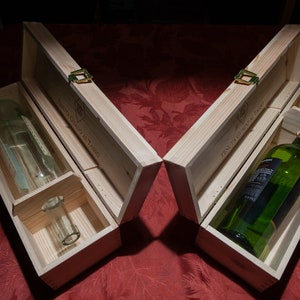 May include: Two light-colored wooden boxes with hinged lids and metal latches. One box contains a clear glass bottle and a small glass piece, while the other holds a green glass bottle. The text Tenuta Guado al Tasso is engraved on the lids.