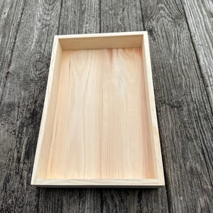 Puede incluir: Una bandeja rectangular de madera clara con un acabado natural. La bandeja tiene un diseño simple y limpio con un borde elevado. La veta de la madera es visible, agregando textura. La bandeja está vacía.