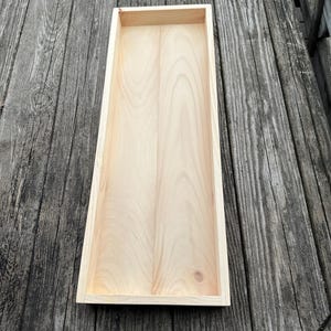 Puede incluir: Una bandeja rectangular de madera con un acabado de madera natural. La bandeja tiene un borde elevado y está hecha de madera sin tratar.